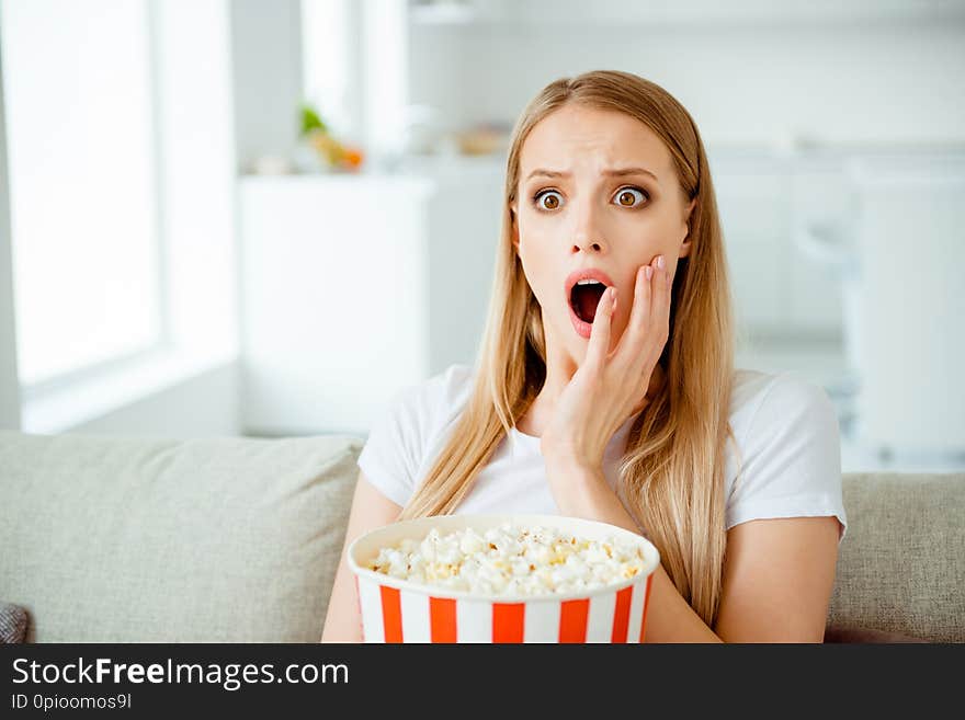 Close up photo of attractive nice student youth impressed astonished watch see incredible tv program entertainment scream shout touch cheeks sit in light apartment dressed trendy modern t-shirt.