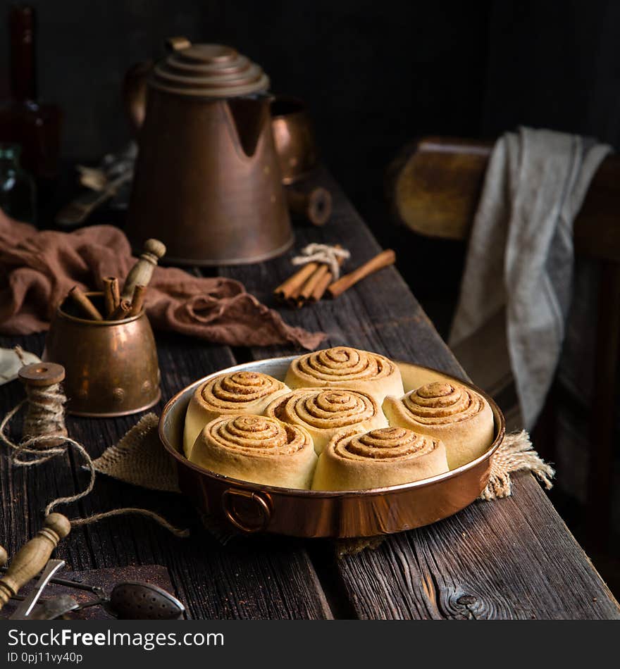 Homemade cinnamon roll buns in vintage copper round pan on wooden rustic table with teapot, cups, cinnamon sticks, towel, strainer. Cinnabon. Dark foodphoto