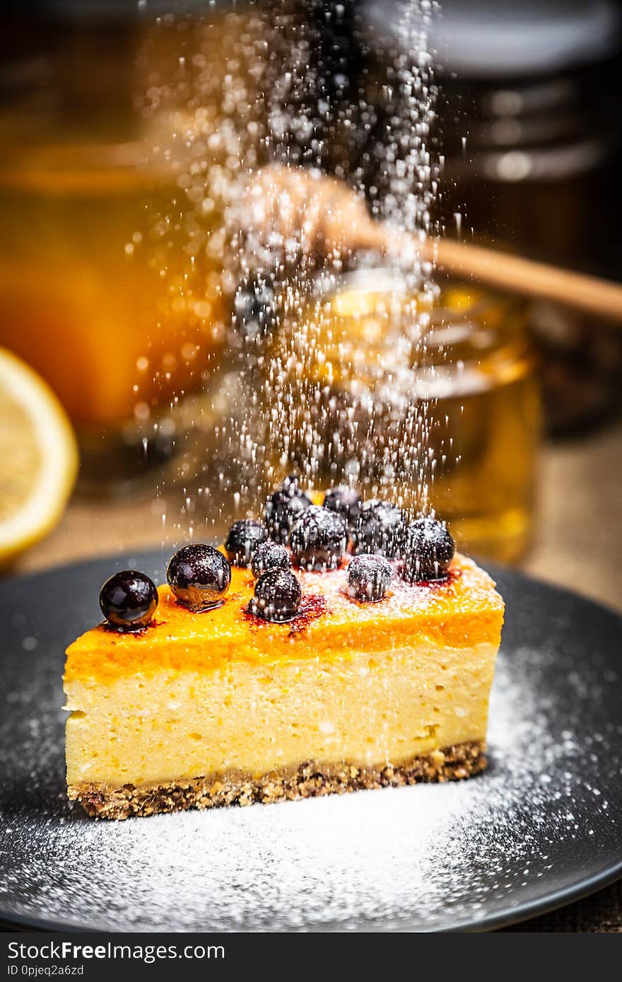 Slice of cheesecake with berries on a blue plate with honey and lemon slices. Blurred background, rural style. Sugar powder. Slice of cheesecake with berries on a blue plate with honey and lemon slices. Blurred background, rural style. Sugar powder