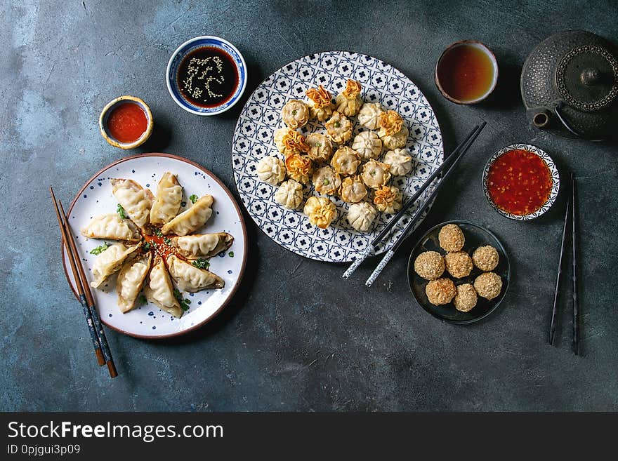 Dim sum Gyozas asian fried dumplings party set with variety of sauces served in ceramic plates and bowls with chopsticks, tea cups and teapot over dark blue texture background. Flat lay, space. Dim sum Gyozas asian fried dumplings party set with variety of sauces served in ceramic plates and bowls with chopsticks, tea cups and teapot over dark blue texture background. Flat lay, space
