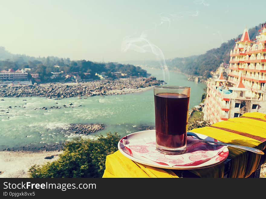 A glass of coffee on a plate against the background of the beautiful scenery of Rishikesh.. View of Ganga river embankment, Lakshman Jhula bridge and Tera Manzil Temple, Trimbakeshwar in Rishikesh