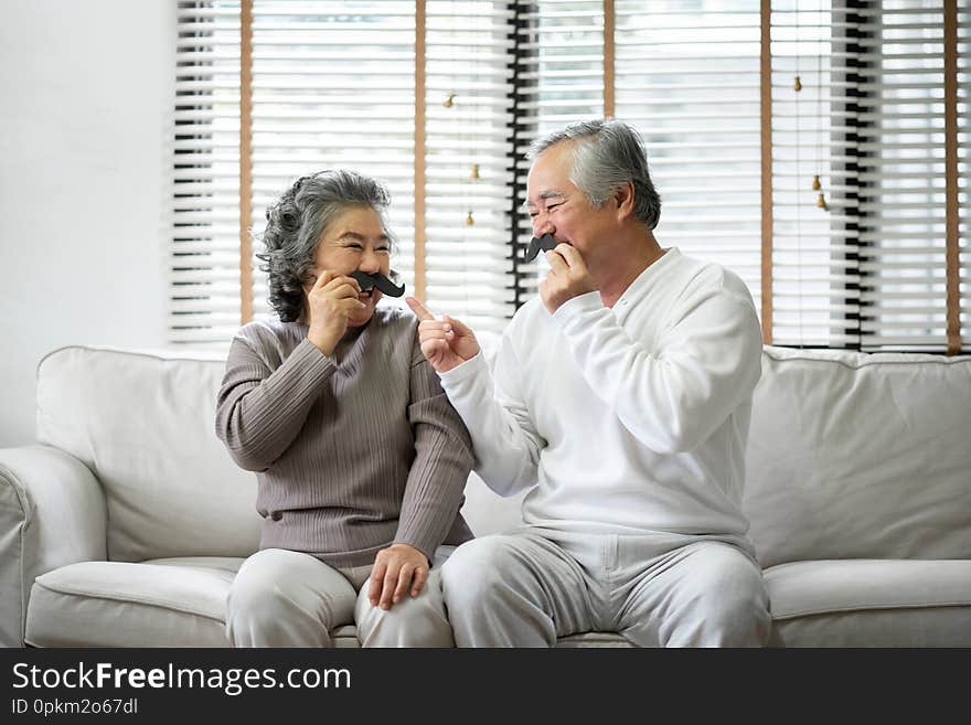 Asian Senior Couple are sitting on sofa and playing paper mustaches at home togetherness. Copy space