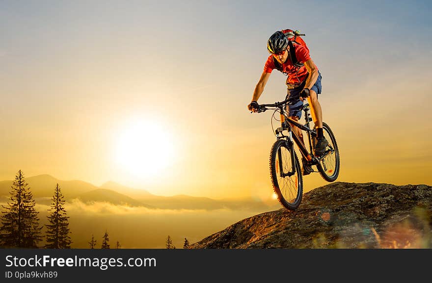 Cyclist in Red T-Shirt Riding the Bike in the Beautiful Mountains Down the Rock on the Sunrise Sky Background. Extreme Sport and Enduro Biking Concept. Cyclist in Red T-Shirt Riding the Bike in the Beautiful Mountains Down the Rock on the Sunrise Sky Background. Extreme Sport and Enduro Biking Concept.