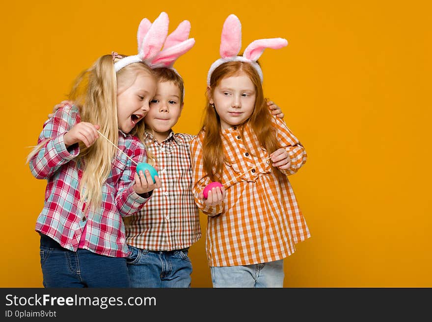 Two little girls and boy with Easter bunny ears holding colorful eggs on yellow background. Happy easter. cute children