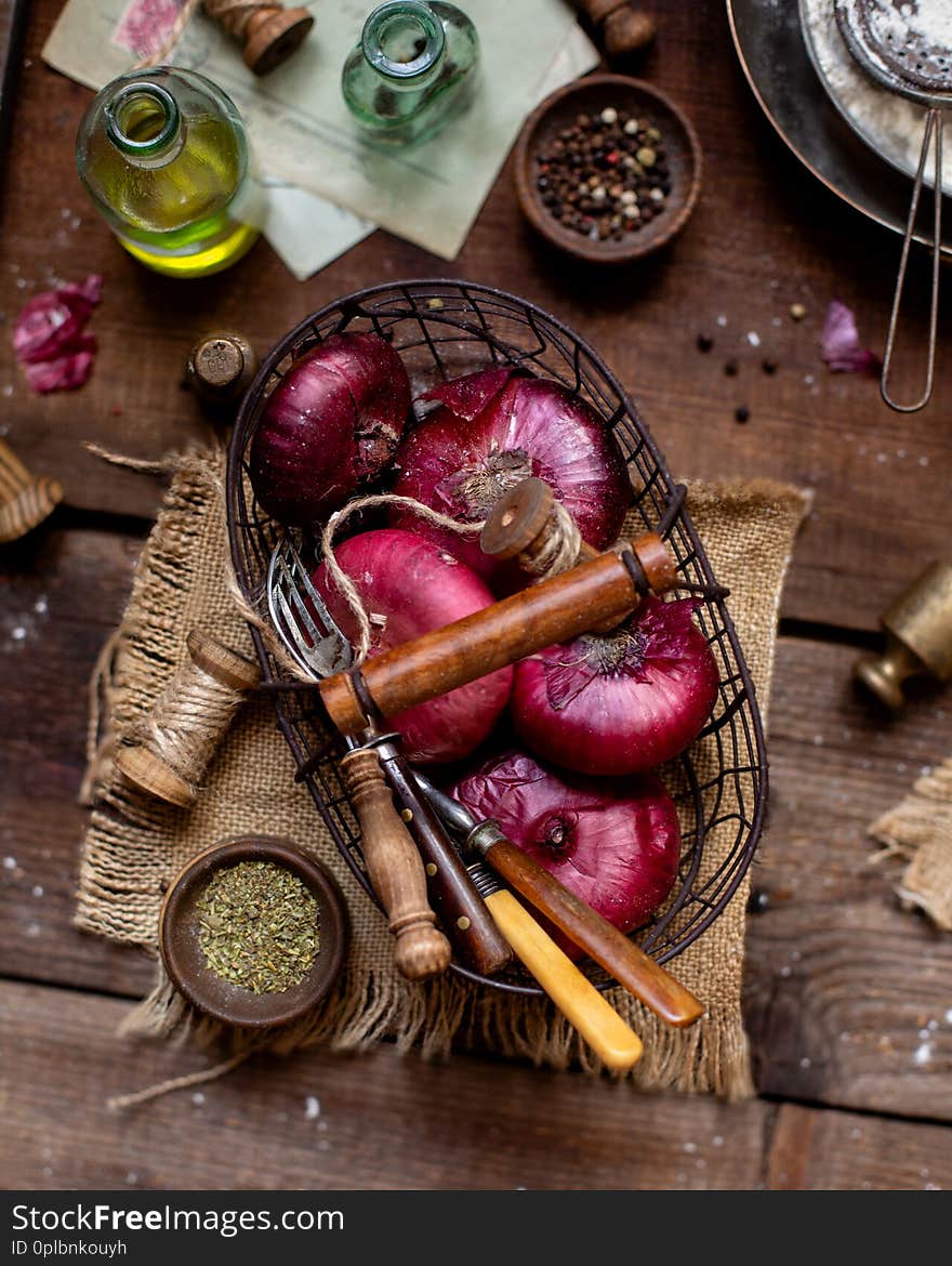 Overhead shot of metal basket of red onion, spoons, strings standing on wooden brown table with sackcloth, bottle with oil, spices, pepper, plate with flour, vintage weights. Ingredients for cooking. Overhead shot of metal basket of red onion, spoons, strings standing on wooden brown table with sackcloth, bottle with oil, spices, pepper, plate with flour, vintage weights. Ingredients for cooking