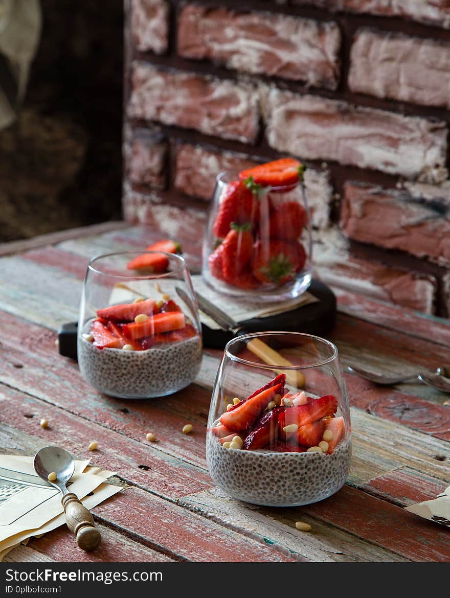 Two glasses with homemade super food chia pudding with cut strawberries on red rustic wooden table with glass of berries, knife, seeds opposite red brick wall. Breakfast still life