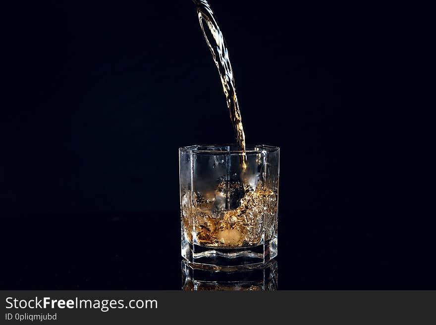 Whiskey pouring into glass with ice isolated on reflective black surface. Whiskey splash out of glass, many drops of beverage get out from glass. Whiskey pouring into glass with ice isolated on reflective black surface. Whiskey splash out of glass, many drops of beverage get out from glass