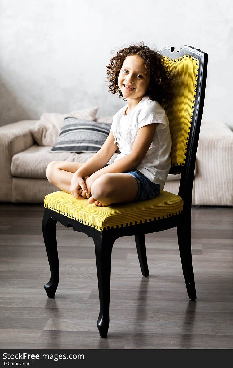 A pretty cute little girl with curly hair sitting on the chair and smiling.