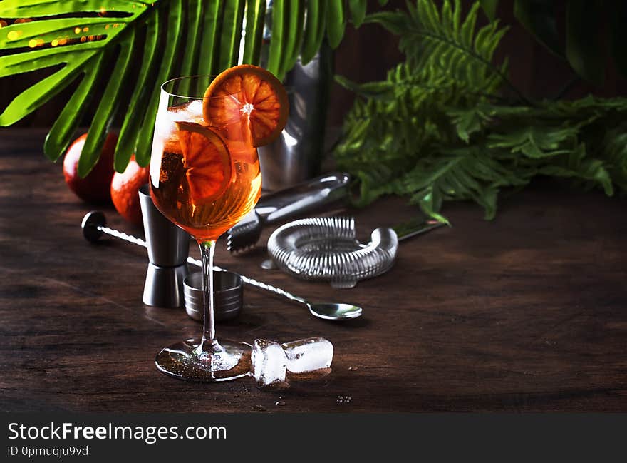 Aperol spritz cocktail in big wine glass with orange slices, summer cool fresh alcoholic cold beverage. Wooden bar counter background with tools, summer mood concept with palm leaves, copy space, selective focus