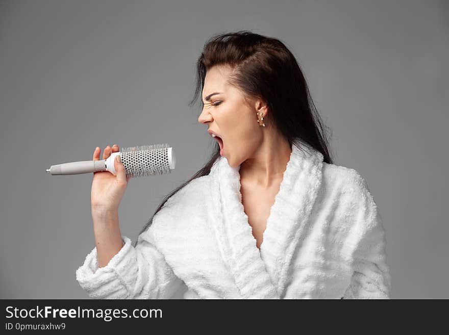 A beautiful brunette in a white terry robe with disheveled hair sings emotionally using a comb instead of a microphone. Studio photo on a gray background. A woman has a good mood in the morning. The girl is going to the party.