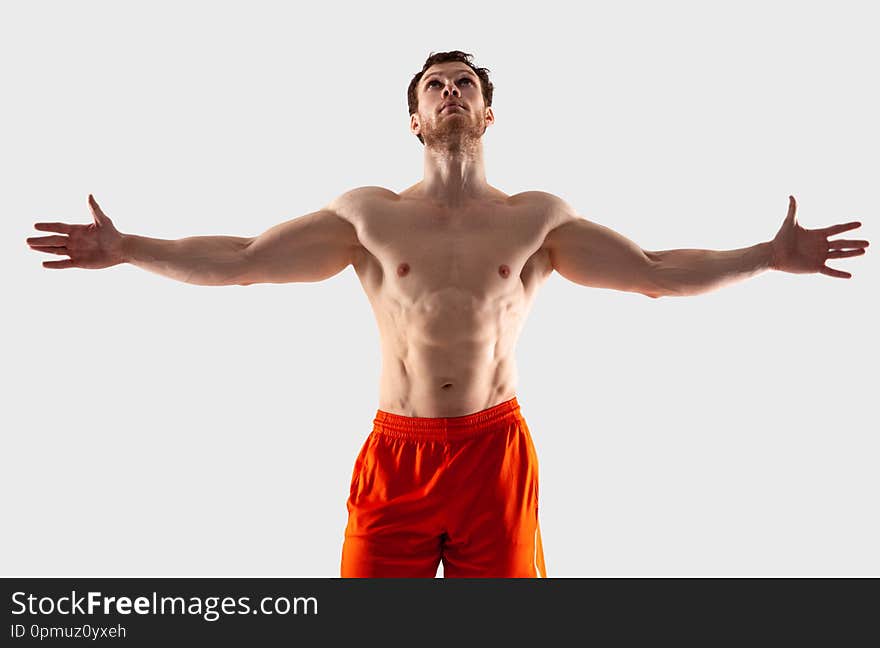 A man in orange shorts with a muscular body spread his arms to the side and looks up