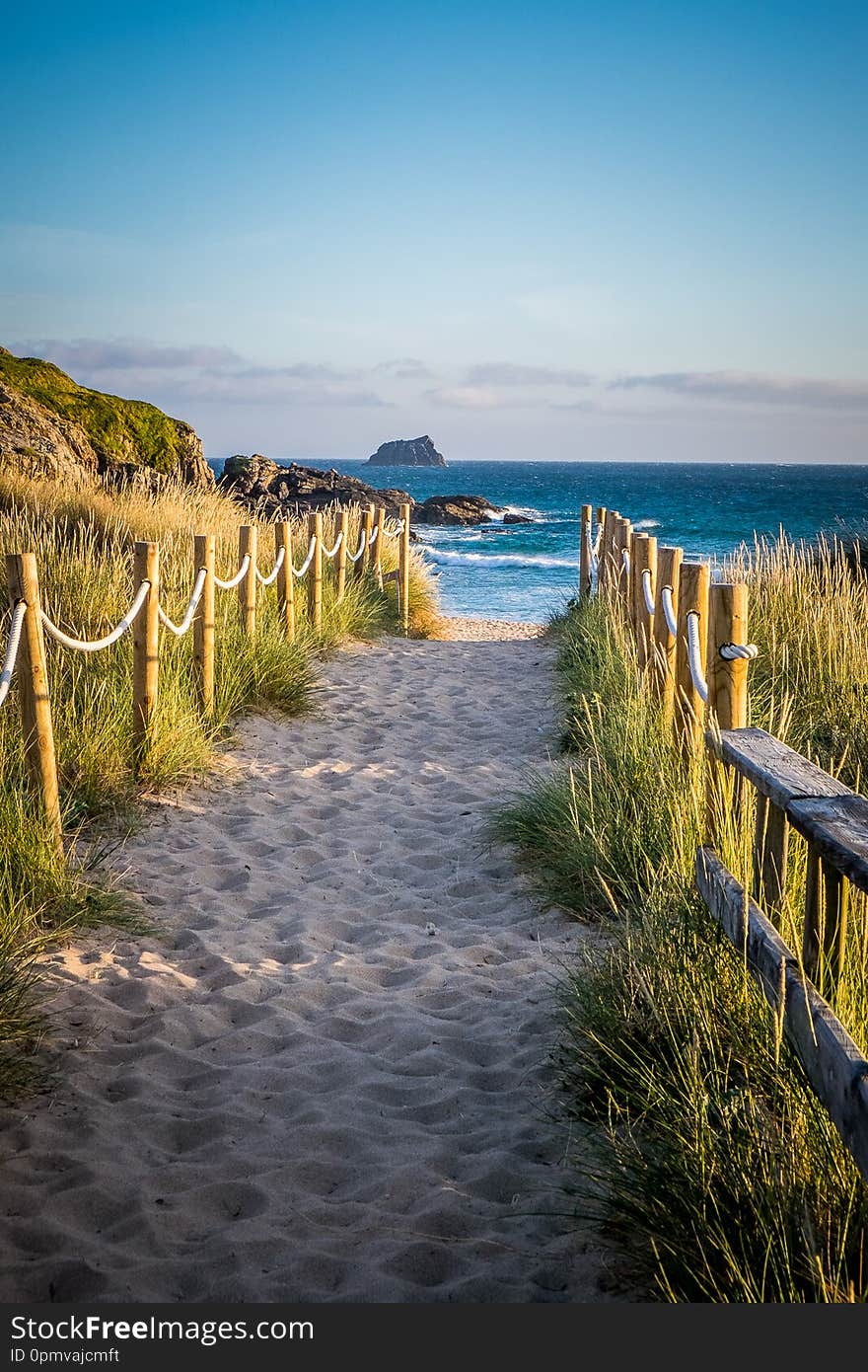 Photograph of a sandy well trodden path to the beach in Finesterre Spain. There is a rope and stick fence on each side with grasses beyond. Blue water and blue sky in the background. Photograph of a sandy well trodden path to the beach in Finesterre Spain. There is a rope and stick fence on each side with grasses beyond. Blue water and blue sky in the background.