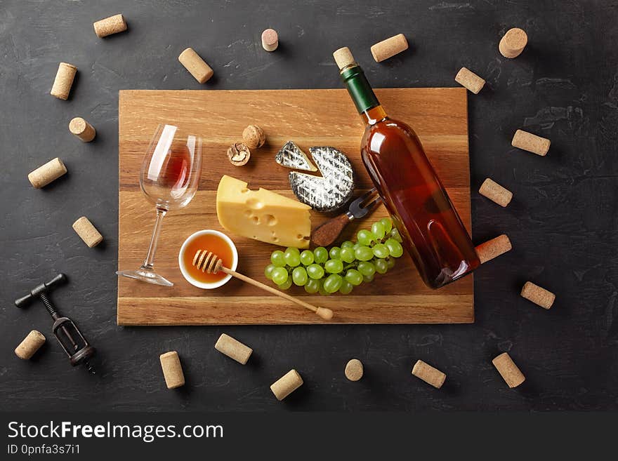 White wine bottle, cheese head, bunch of grapes, honey, nuts and wineglass on cutting board with corks and corkscrew on black background. Top view with copy space