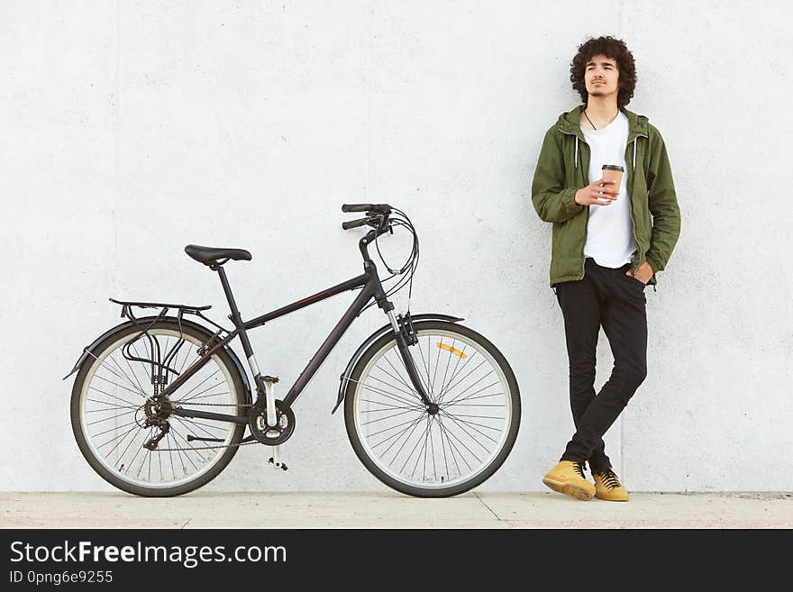 Indoor shot of attractive pencive man with crisp hair drinking hot baverage from paper cup, looks thoughtfully away, wears in fashionable clothes, stands near bicycle, isolated over white studio wall