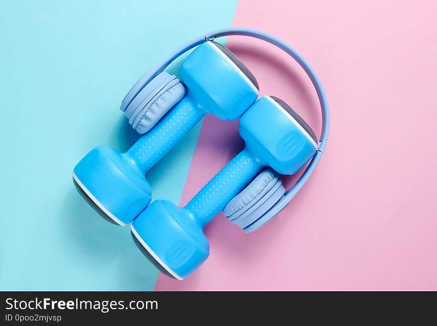 Minimalism. Blue headphones, plastic dumbbells on blue pink background. Sports with music. Flat lay. Top view