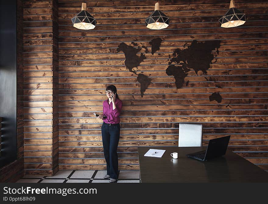 Businesswoman standing chatting on her mobile phone in a large spacious office below a decorative map of the world on the wall