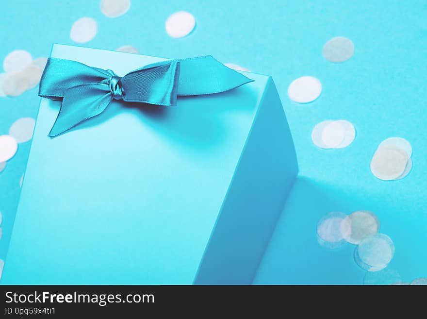 Fragment of blue box with shiny satin ribbon tied in a bow with scattered pastel confetti on the background of the same tone. Trendy creative celebration backdrop with copy space for text. Fragment of blue box with shiny satin ribbon tied in a bow with scattered pastel confetti on the background of the same tone. Trendy creative celebration backdrop with copy space for text