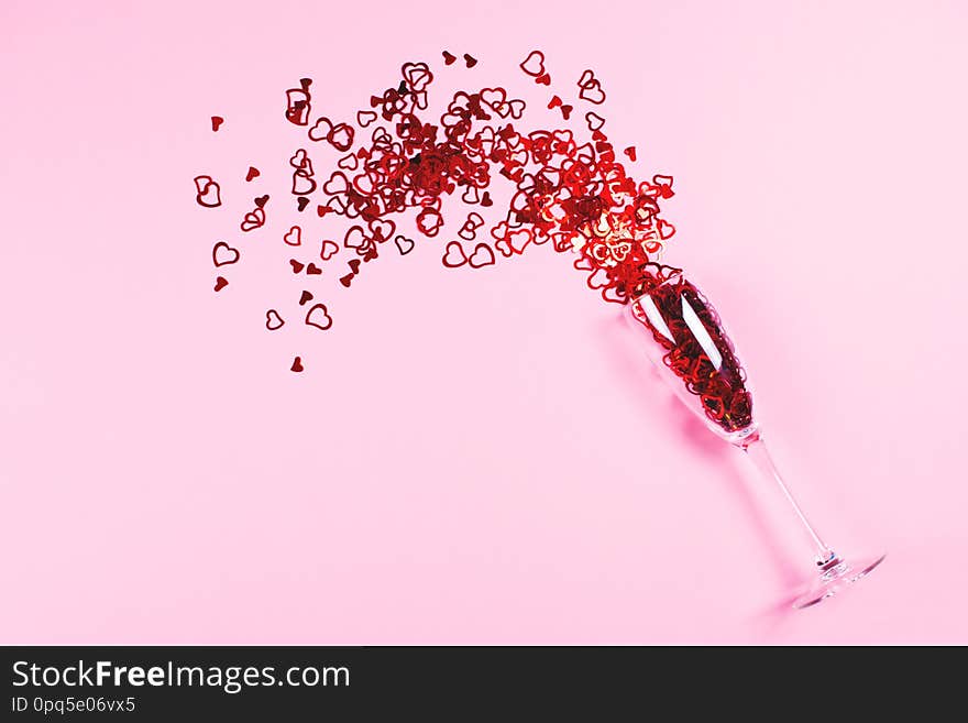 Heart-shaped vibrant red confetti poured out of champagne glass on pink background with copy space for text. Top view and flat lay style. Holiday, love and celebration concept. Heart-shaped vibrant red confetti poured out of champagne glass on pink background with copy space for text. Top view and flat lay style. Holiday, love and celebration concept