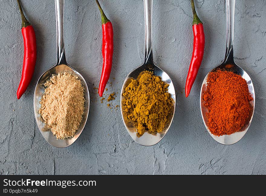 Different spices in metall spoons on grey background. Red pepper. Ginger root. Carnation. Pepper pods. Cinnamon. Macro. Selective focus. Close up. Side view. Different spices in metall spoons on grey background. Red pepper. Ginger root. Carnation. Pepper pods. Cinnamon. Macro. Selective focus. Close up. Side view.