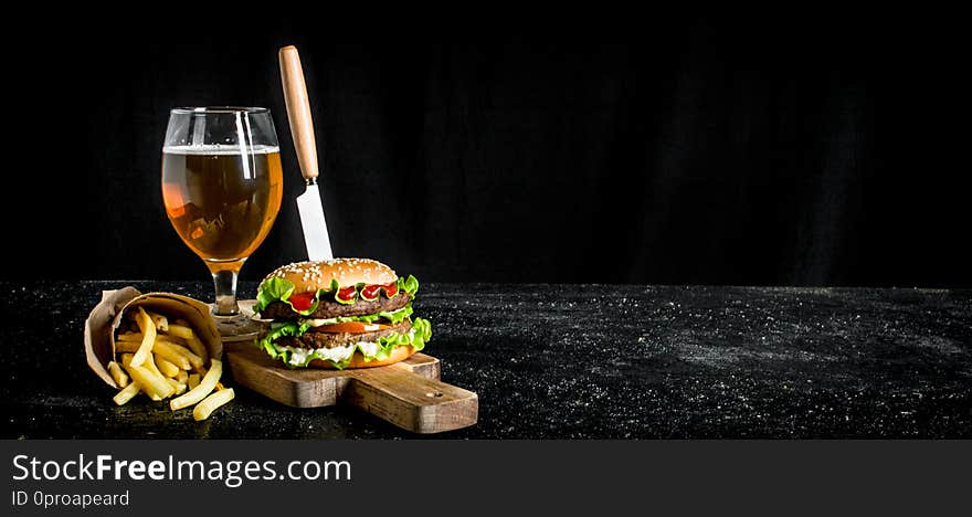 Burger with a knife,fries, beer in a glass. On black rustic background