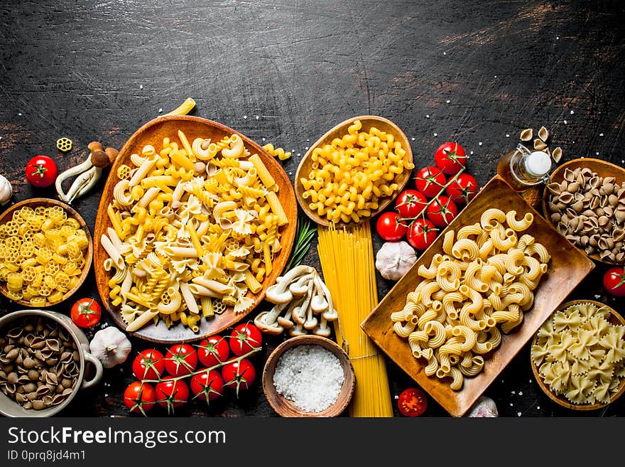 Assortment of different types of raw pasta with mushrooms, tomatoes and garlic. On dark rustic background