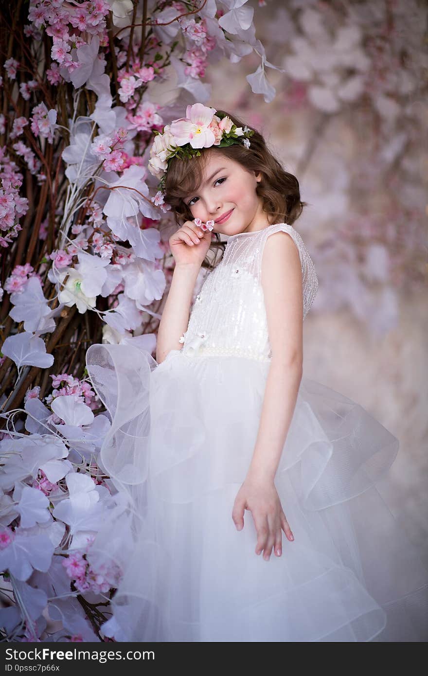 A child in a beautiful long white dress with a flower wreath on his head. On the background of white and pink flowers. Studio shooting. A child in a beautiful long white dress with a flower wreath on his head. On the background of white and pink flowers. Studio shooting.