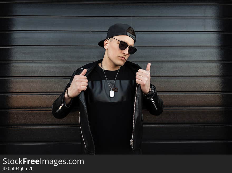 Serious young hipster man in a black baseball cap in stylish dark sunglasses in a vintage leather jacket with a fashionable t-shirt stands near a black wooden house. Handsome urban guy outdoors