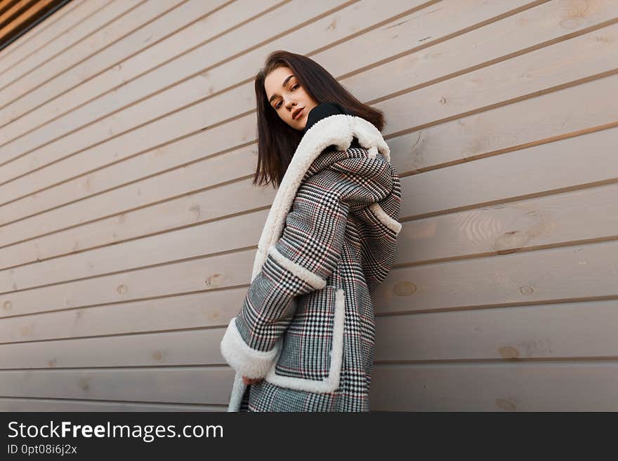 Young pretty attractive woman in a luxurious checkered jacket  with white fur in retro style is posing on the street near a vintage wooden house. Fashionable beautiful girl fashion model. Youth style