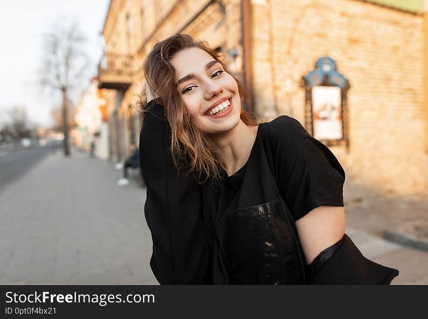Young joyful beautiful woman with a wonderful smile in a stylish T-shirt in a elegant coat posing on a sunny day in the city near vintage brick buildings. Positive cheerful girl on a walk. Spring