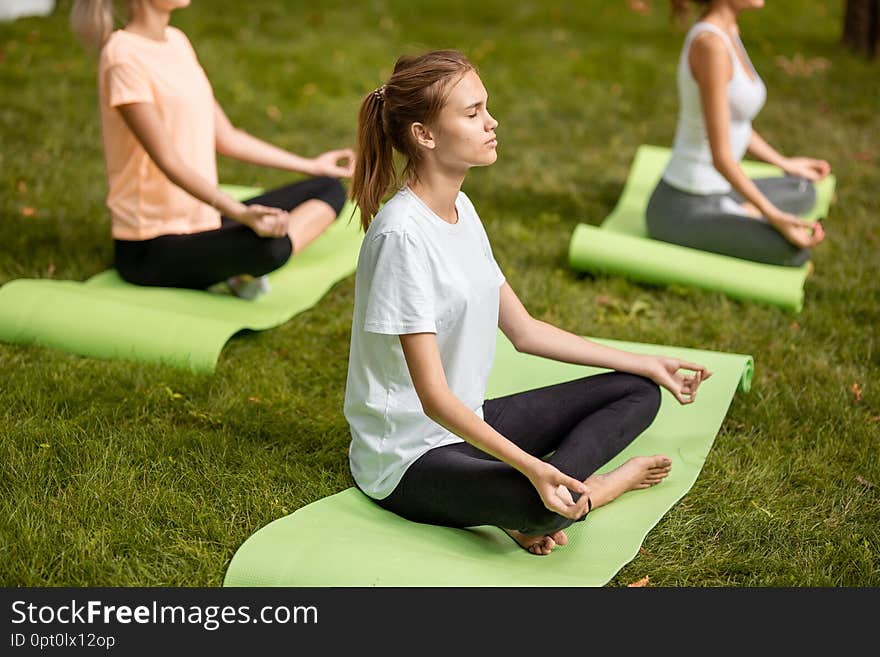 Three young slim girls sit in the lotus positions with closing eyes doing yoga on yoga mats on green grass in the park