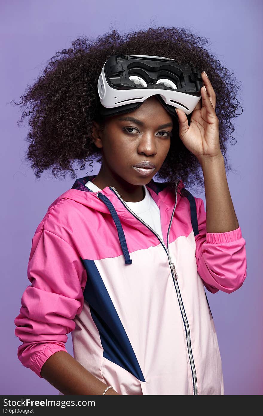 Mod curly brown-haired girl dressed in the pink sports jacket wearing on her head the virtual reality glasses poses in the studio on neon background .