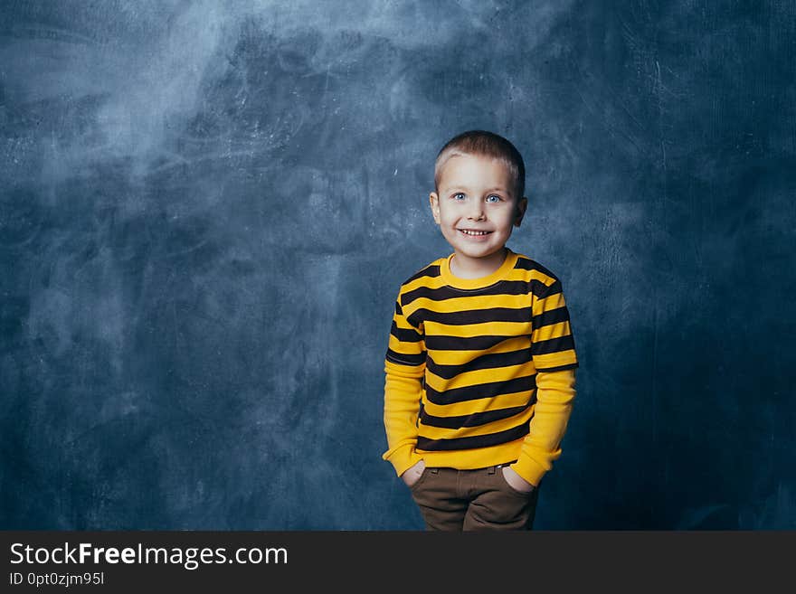 A little boy poses in front of a gray-blue concrete wall. Portrait of a smiling child dressed in a black and yellow striped