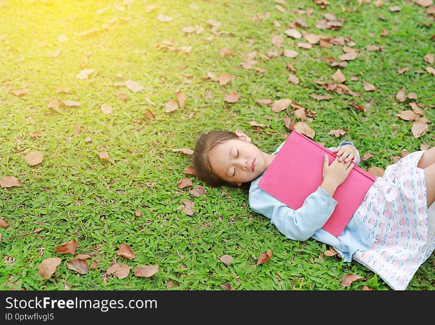 Little girl resting with book lying on green grass with dried leaves in the summer garden.
