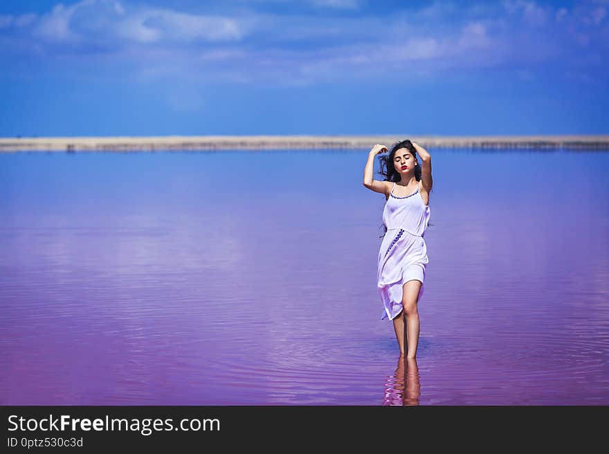 Beautiful young girl in long white dress posing on salty pink lake.