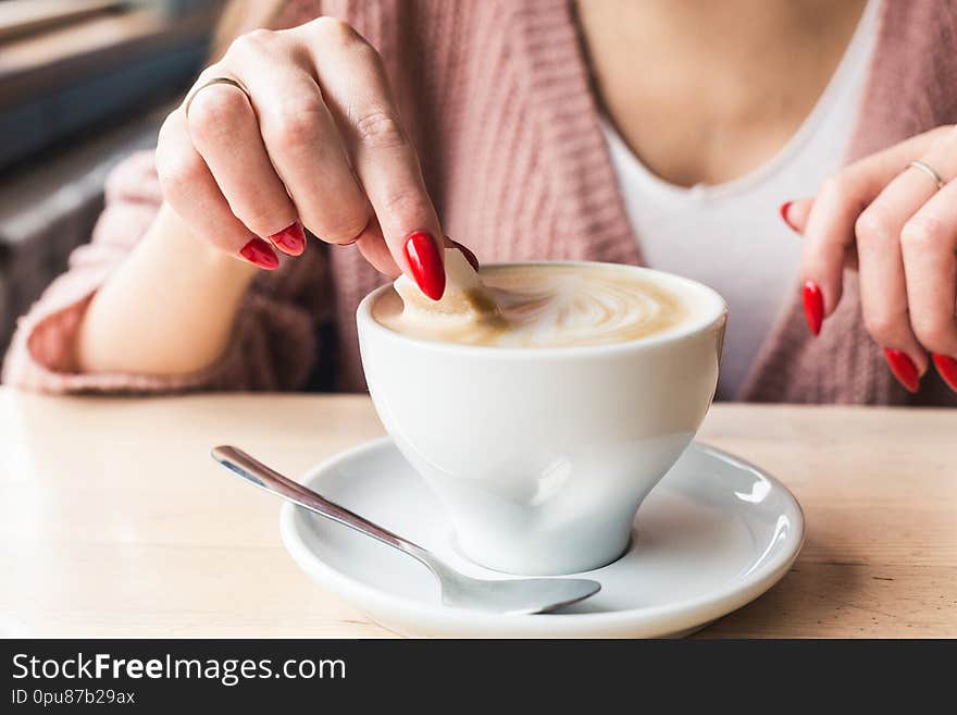 A girl dunks a piece of sugar in coffee. Closeup of hands with red nails make-up and cappuccino cup