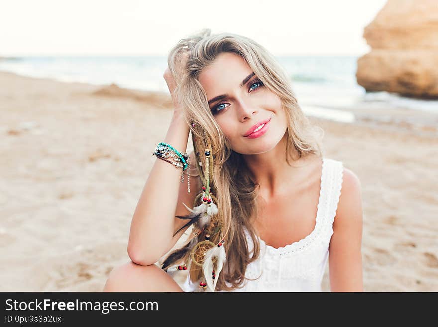 Closeup portrait of attractive blonde girl with long hair and blue eyes sitting on  beach. She wears  white dress, ornamentation. She is looking to the camera. Closeup portrait of attractive blonde girl with long hair and blue eyes sitting on  beach. She wears  white dress, ornamentation. She is looking to the camera.