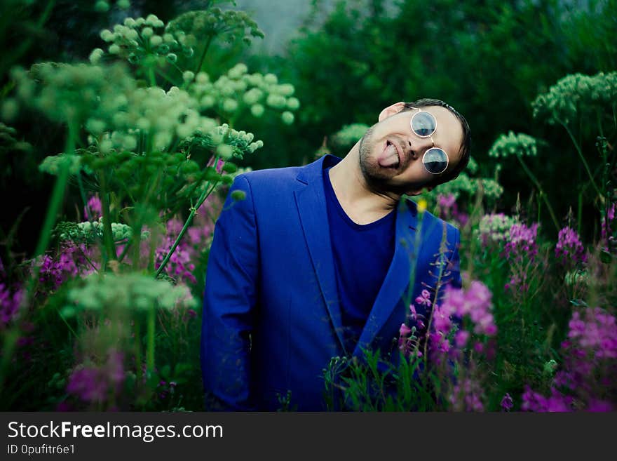A man in a blue jacket on a flower meadow. He`s wearing blue glasses. In the meadow of purple flowers and large hats of white high flowers.