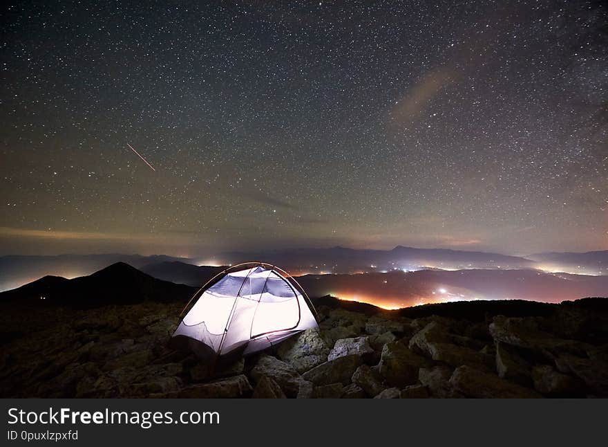 Tourist summer camping at night on the top of rocky mountain. Glowing tent under incredibly beautiful night sky full of stars. On background amazing starry sky, mountains and luminous villages. Tourist summer camping at night on the top of rocky mountain. Glowing tent under incredibly beautiful night sky full of stars. On background amazing starry sky, mountains and luminous villages