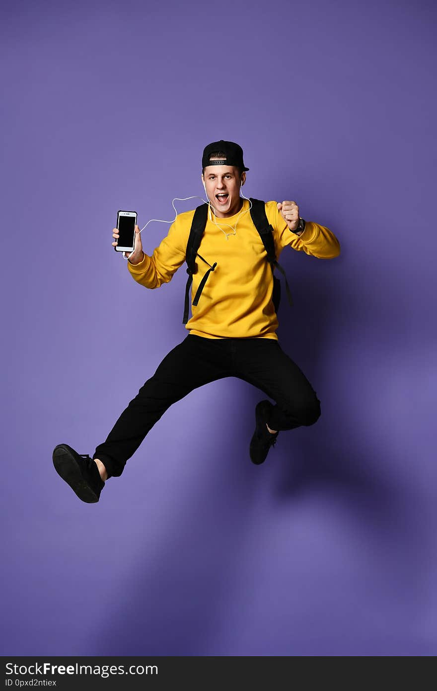 Handsome young student man in casual wear with backpack and headphones is listening to music using a smartphone and smiling while jumping on purple background