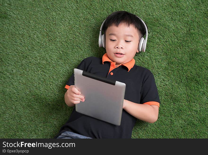 Top view of little boy in headphones using a digital tablet and smiling while lying on green grass.