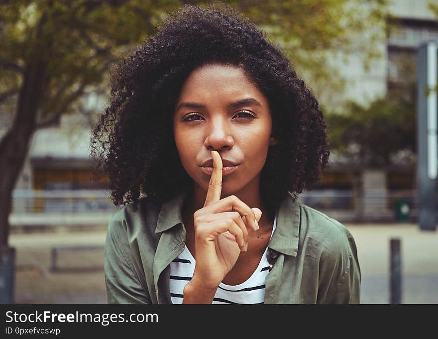 Portrait of a young woman looking at camera with finger on her lips making the silence sign in the city. Portrait of a young woman looking at camera with finger on her lips making the silence sign in the city