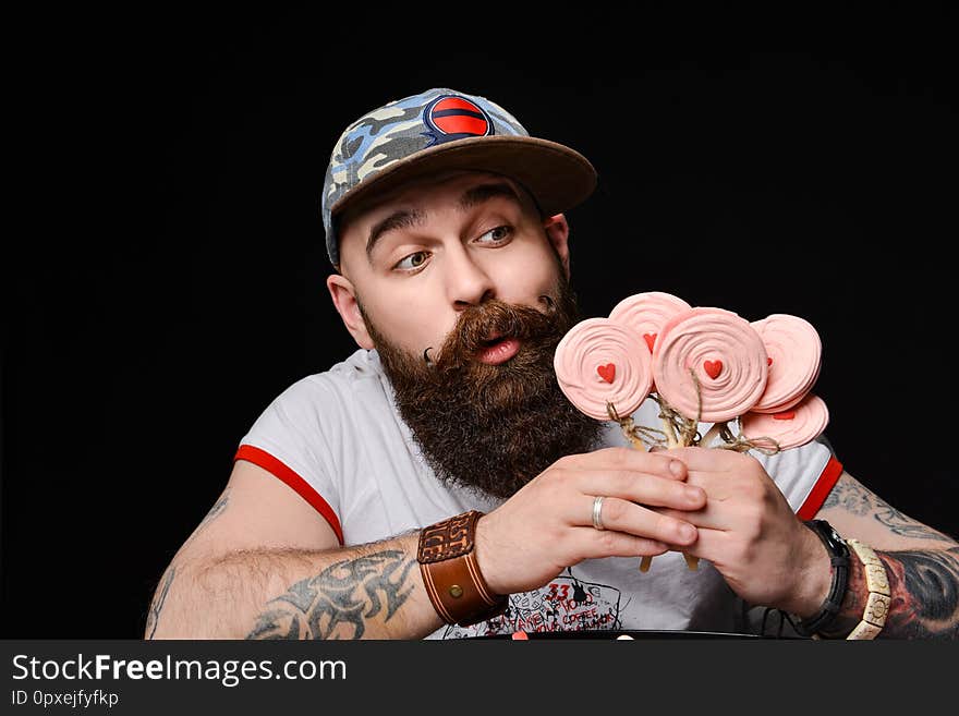 Happy Bearded Brutal Man In A Fashionable Cap Holds A Pack Of Lollipops Candies