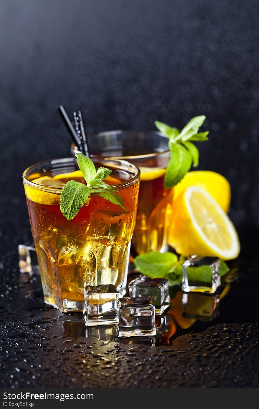 Two glasses with cold traditional iced tea with lemon, mint leaves and ice cubes on wet black background