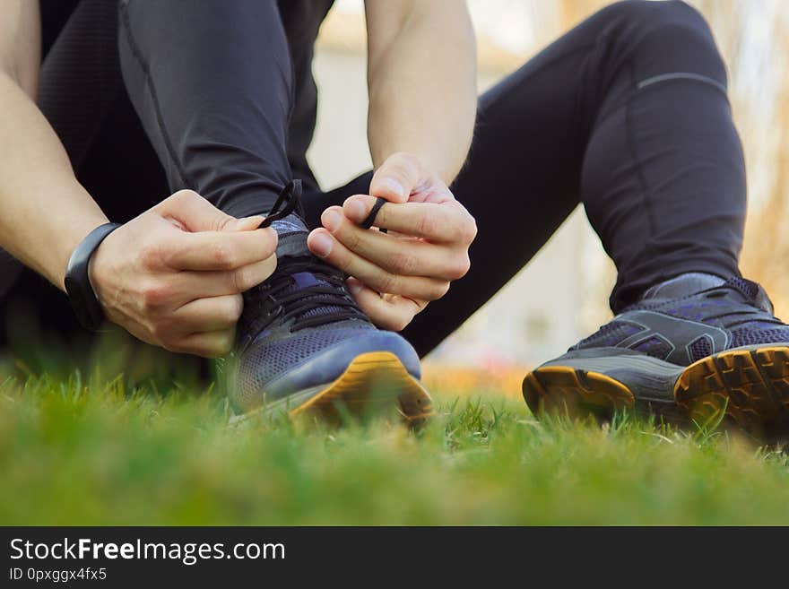 A young man in black clothes is tying the laces on the sneakers close up. fitness athlete sitting on the sports field on the grass. warm up body preparation for the training