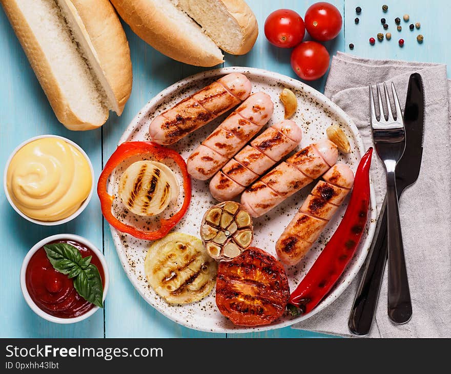 Top view of chicken homemade sausages, buns bread, sauces ketchup and mustard on blue wooden background. Grilled sausages and grilled vegetables in craft trendy plate. Homemade hotdogs. Top view of chicken homemade sausages, buns bread, sauces ketchup and mustard on blue wooden background. Grilled sausages and grilled vegetables in craft trendy plate. Homemade hotdogs.