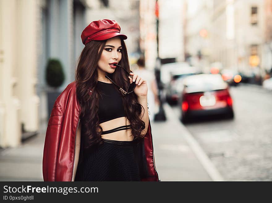 Beautiful fashionable sexy woman with makeup posing on city street wearing a red jacket, hat and sunglasses.