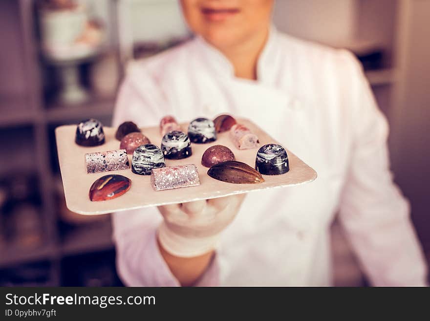 Life enjoyment. Positive delighted woman working in confectionery and holding pralines on the plate. Life enjoyment. Positive delighted woman working in confectionery and holding pralines on the plate