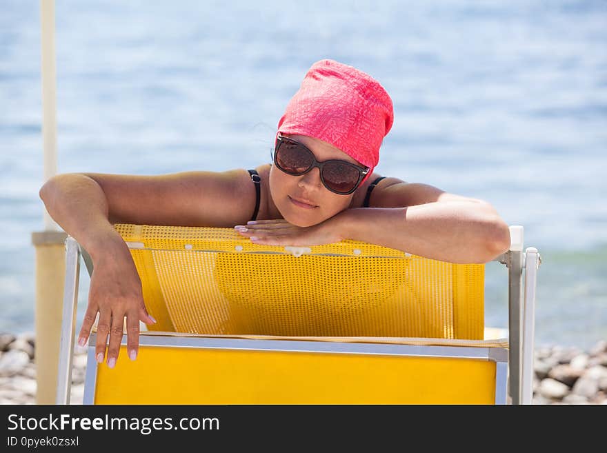 Pleased Caucasian woman in sunglasses sunbathing on yellow sun lounge on beach, looking at camera