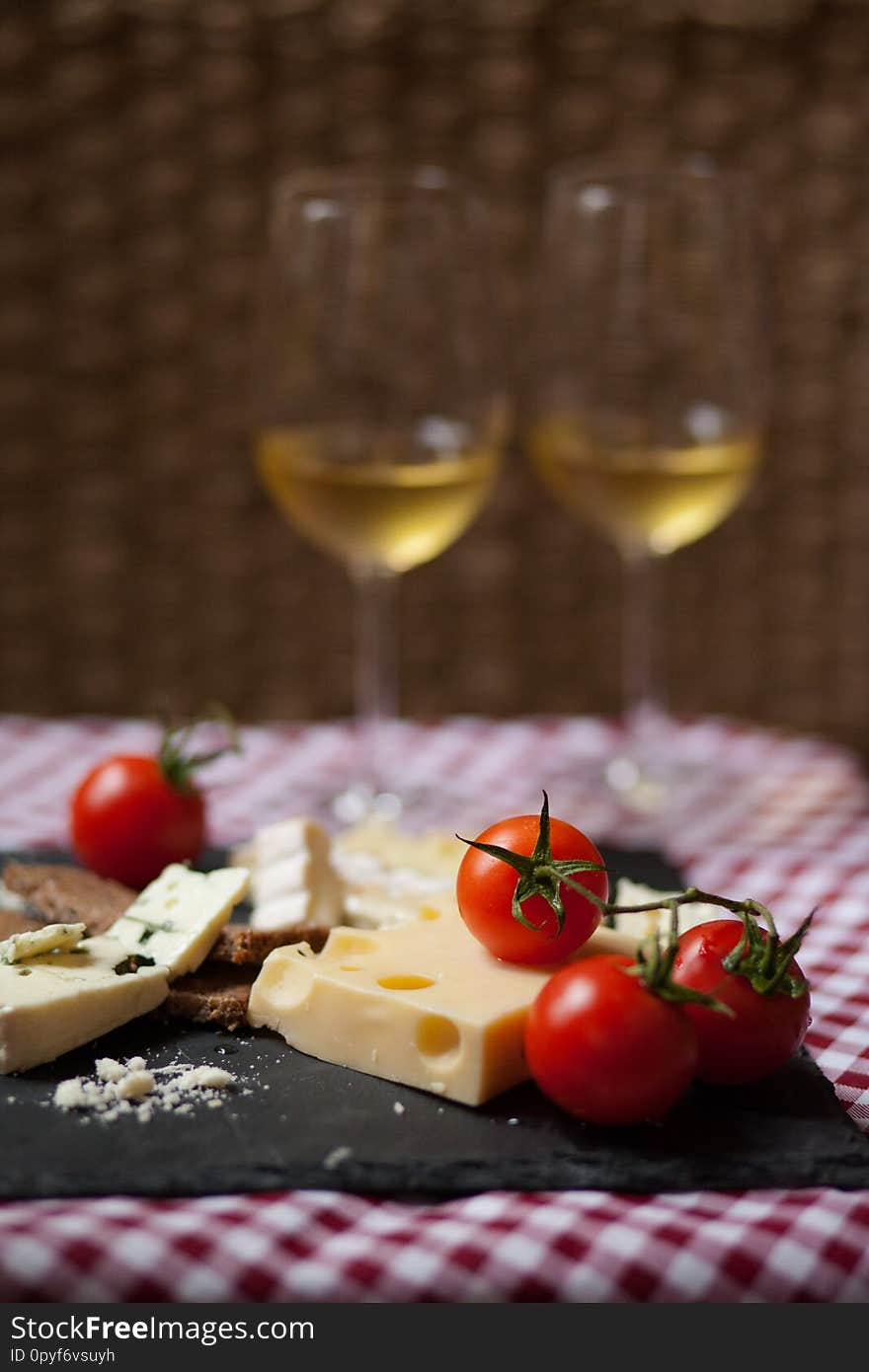 Delicious plate of french cheese served with fresh ripe tomatoes and bread on a black charcoal board. Red and white tablecloth for a picnic mood. Two glasses of white wine on background. Delicious plate of french cheese served with fresh ripe tomatoes and bread on a black charcoal board. Red and white tablecloth for a picnic mood. Two glasses of white wine on background