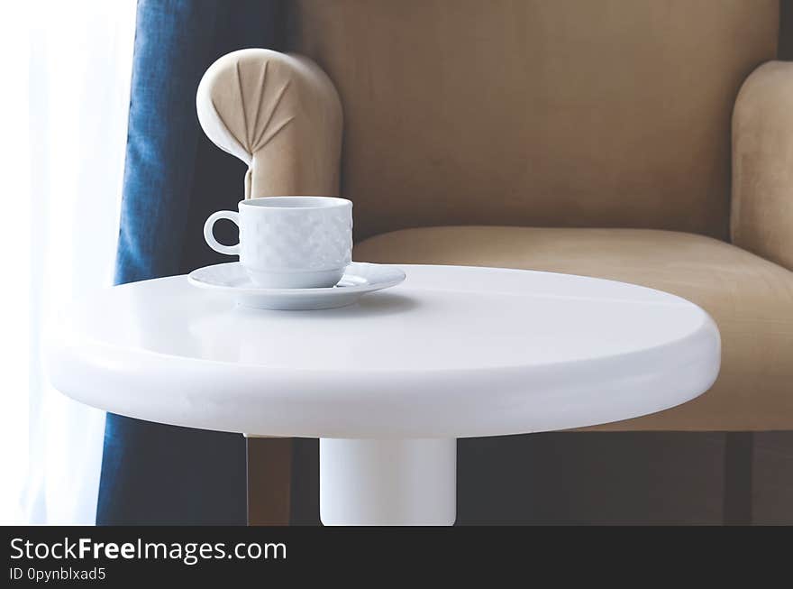 White tea cup and saucer on a round white table against a beige armchair